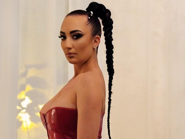 MiaBlackXO mit einem langen, schwarzen Zopf, trägt ein glänzendes, rotes Top ohne Träger, das ihre Brüste zeigt, hat auffälliges Make-up und einen ernsten Gesichtsausdruck..