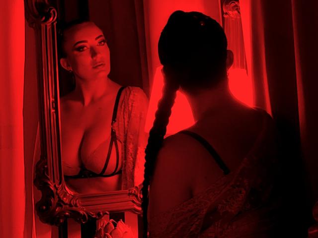 MiaBlackXO stands in front of a mirror, red lighting highlighting her large tits in a black lace bra, looking at herself with a serious expression, dark braids over shoulder.