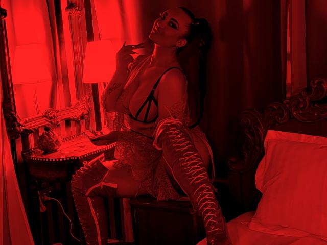 MiaBlackXO com tatuagens, vestindo lingerie de renda e botas de renda até a coxa, sentada em uma cama de madeira escura, luz vermelha, pose sedutora, mão perto da boca, rabo de cavalo comprido, quarto..