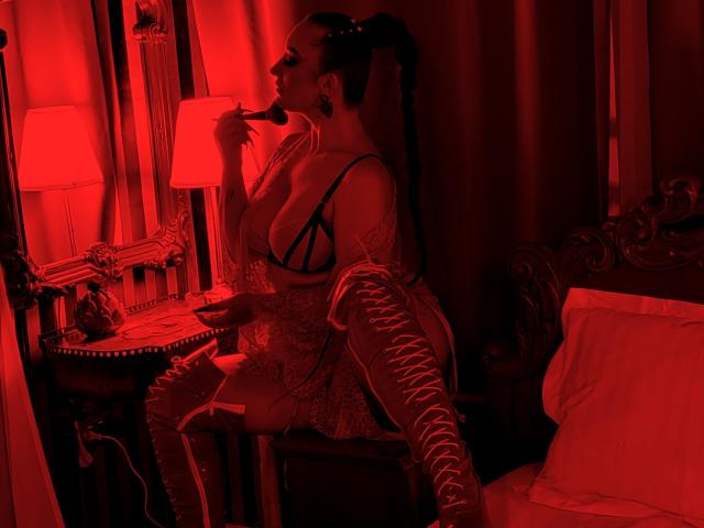 MiaBlackXO sits in red-lit room, wearing lace bra, sheer top, and long lace-up gloves, with high ponytail, touching her chin, showing her tits, in ornate bedroom.