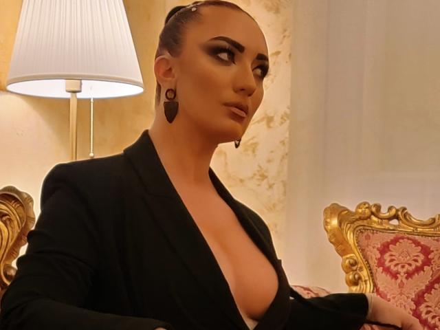 MiaBlackXO con i capelli scuri raccolti in uno chignon, indossa un blazer nero, mostrando il suo seno prosperoso con una camicia a scollo profondo, orecchini neri e una sedia rossa con bordi dorati..