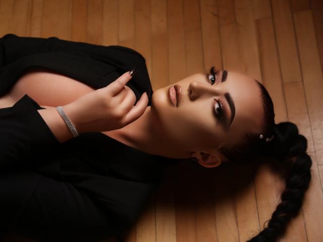 MiaBlackXO with dark braids, lying on wooden floor, wearing black top, exposing her tits, touching her chin, looking at camera, dark makeup, silver bracelet.