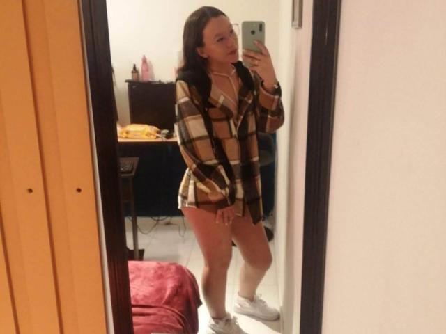 TamyParist a les cheveux bruns, porte une chemise à carreaux, n'a pas de pantalon et des baskets blanches. Elle se prend en photo devant un miroir avec son téléphone, montrant ses fesses et ses petites seins..