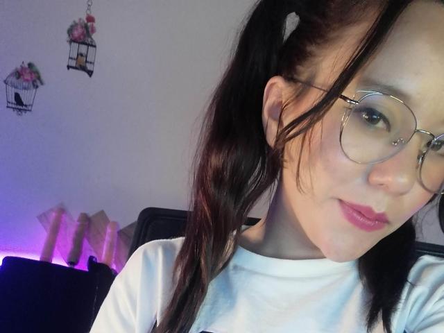 TamyParist, con labios rosados y voluminosos, lleva gafas y una camiseta blanca, tiene trenzas negras, piel clara y una ligera sonrisa, mostrando su rostro y parte superior del pecho..