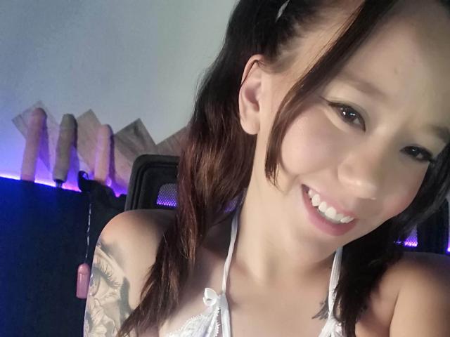 TamyParist con due codini, sorridente, indossa un reggiseno di pizzo bianco, mostra un tatuaggio sul braccio sinistro, ha un leggero tocco rosa sulla guancia e una luce LED viola sullo sfondo. Il seno è visibile attraverso il reggiseno di pizzo..