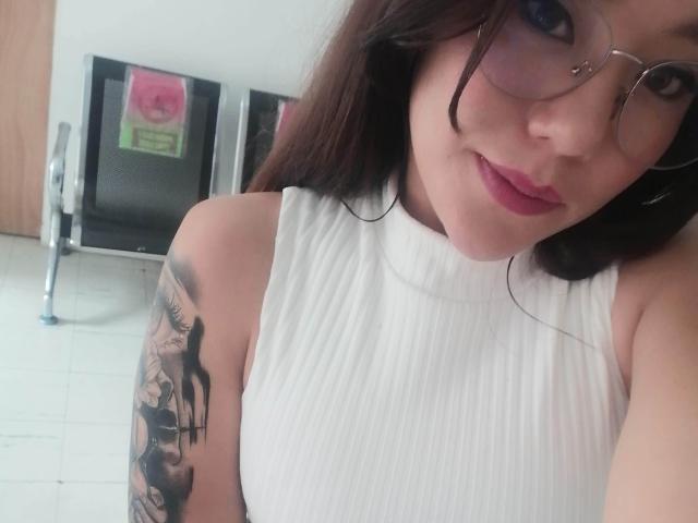 TamyParist with glasses, white sleeveless ribbed top, showing tattooed arm, pink lipstick, and smiling, in a selfie. Boobs not visible. She has dark hair and a cute face.
