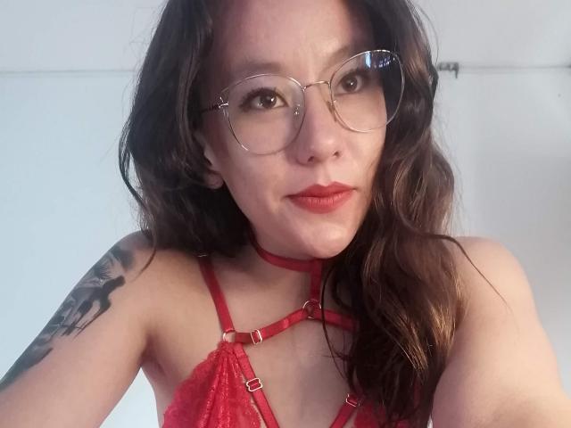TamyParist, com cabelos castanhos ondulados, um sutiã de renda vermelho e óculos, tem uma tatuagem no braço direito e um colar vermelho, exibindo seus seios e batom vermelho..
