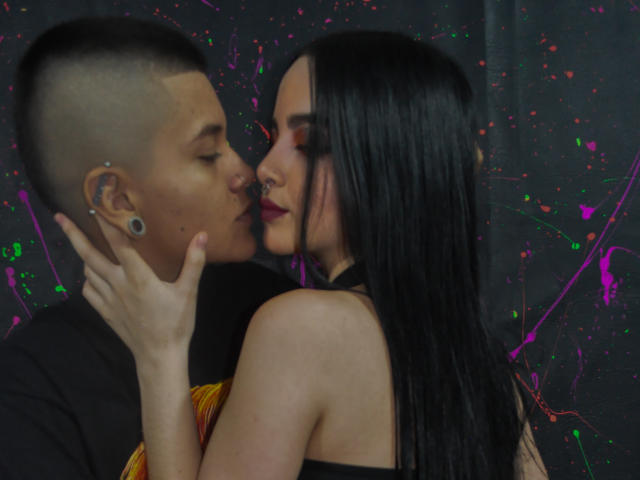 EmilyAndSam, avec des cheveux courts et foncés, des piercings aux oreilles, embrassant EmilyAndSam, avec de longs cheveux noirs, du rouge à lèvres foncé, un haut noir, sur un fond sombre éclaboussé. Sa main est sur sa joue..