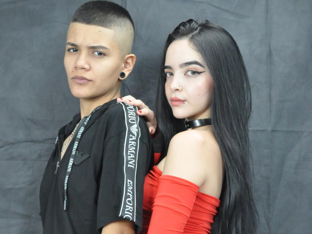 EmilyAndSam, avec des cheveux bruns courts, un t-shirt noir et des bouchons d'oreille, se tient à côté d'EmilyAndSam, avec de longs cheveux noirs, un haut rouge à une épaule et un collier noir, dans une prise de vue en studio..