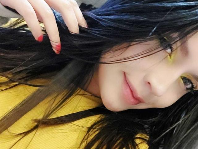 LinaStars with black hair, yellow eyeshadow, red nails, and a slight smile, holding her hair. She's wearing a yellow shirt, and her face is close to the camera.