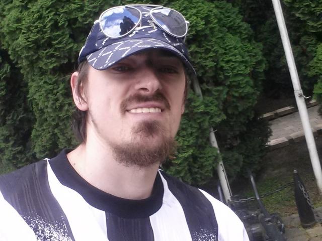 ConanJones has a white and black striped shirt, blue patterned cap with goggles, brown goatee, and is smiling with a slight nose tilt. He's standing outdoors with greenery.
