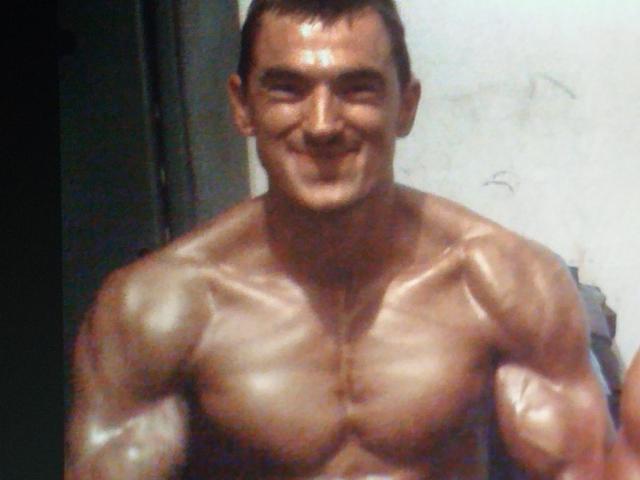 Muscular ConanJones, shirtless with shiny, defined muscles and a broad smile, standing in a dimly lit room. Short dark hair, sweat on his tan skin.