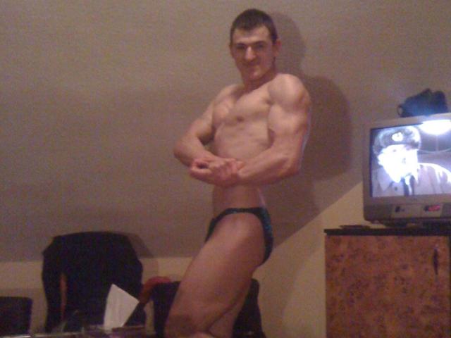 Muscular ConanJones with defined arms and abs, wearing dark blue speedo, standing in room, arms crossed, short brown hair, TV showing blurred image to the right.