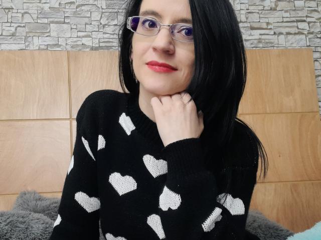 AlanaMoneth with black hair, glasses, red lipstick, black sweater with white heart patterns, resting chin on hand, smiling, pale skin, wooden headboard, gray textured wall behind.