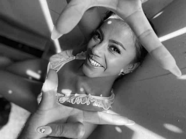 Black-and-white photo of Amberv with a big smile, looking up through hands, wearing a frilled top, showing her tits, with a nose ring and cute earrings.