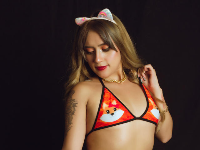Sabrina Cole, con cabello rubio largo, vestida con un top de bikini rojo con motivos de zorros, detalles negros, orejas de gato, una cadena dorada y un tatuaje en el brazo derecho, mirando hacia abajo y sonriendo..