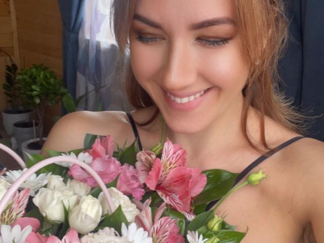 Misstyk with a cute smile, holding a bouquet of pink and white flowers, has light brown hair, and is wearing a black spaghetti strap top, showing her tits.