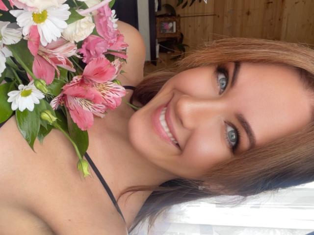 Misstyk with blue eyes, smiling at the camera, holding pink and white flowers, has brown hair, and is wearing a black strap top, showing her tits slightly.