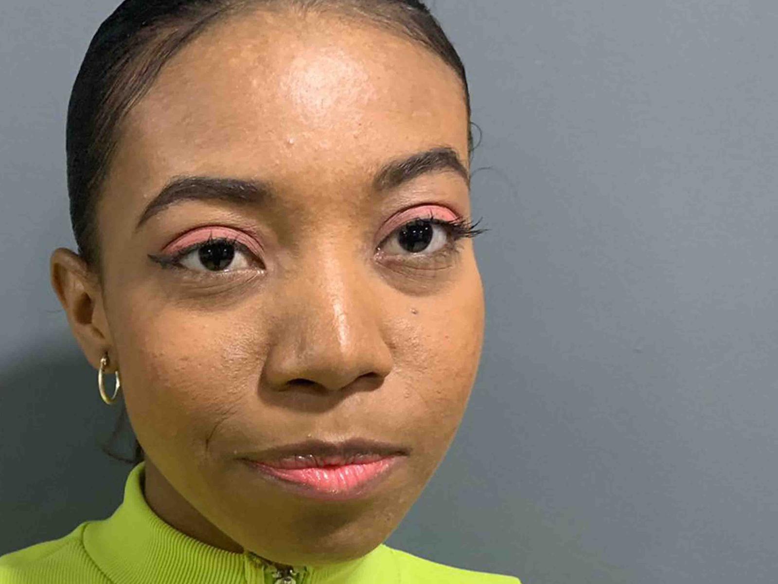Sara Diamante ha una pelle marrone media, ombretto rosa, lunghe ciglia, piccoli orecchini a cerchio e un leggero sorriso con labbra rosate. I suoi capelli sono raccolti e indossa una maglietta verde..