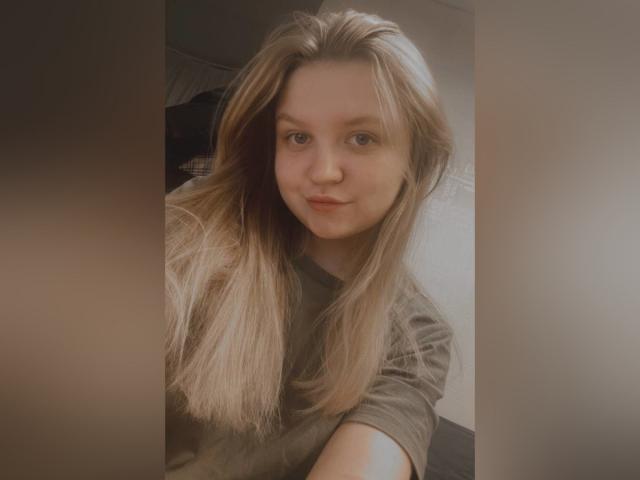 AnitaVine, con lunghi capelli biondi, un sorriso dolce e occhi azzurri, indossa una camicia grigia, in un autoritratto che mette in risalto un trucco leggero e una bellezza naturale..
