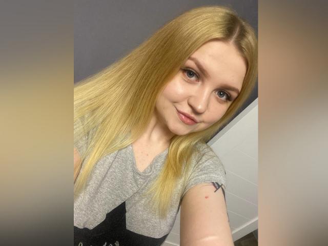 AnitaVine with long blonde hair, small nose, light skin, and a subtle smile, wearing a gray shirt, showing a small tattoo on her right arm.