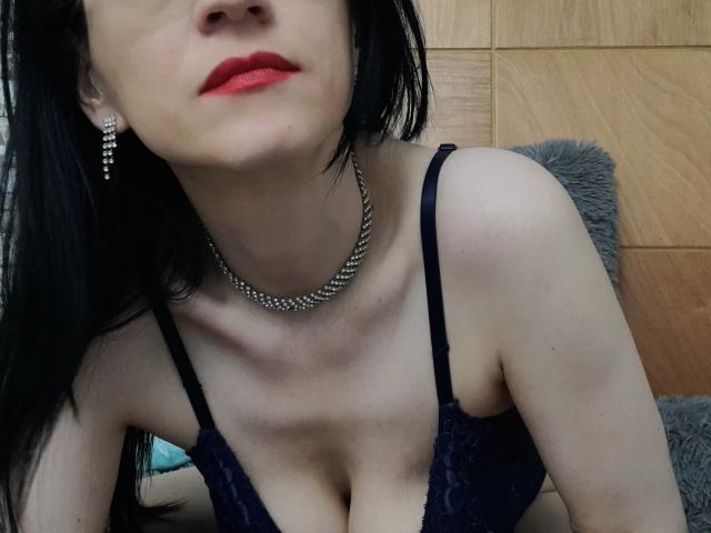 AlanaMoneth with black hair, red lips, wearing a black lace bra, silver necklace, and earrings, showing her tits and collarbone, looking slightly up.