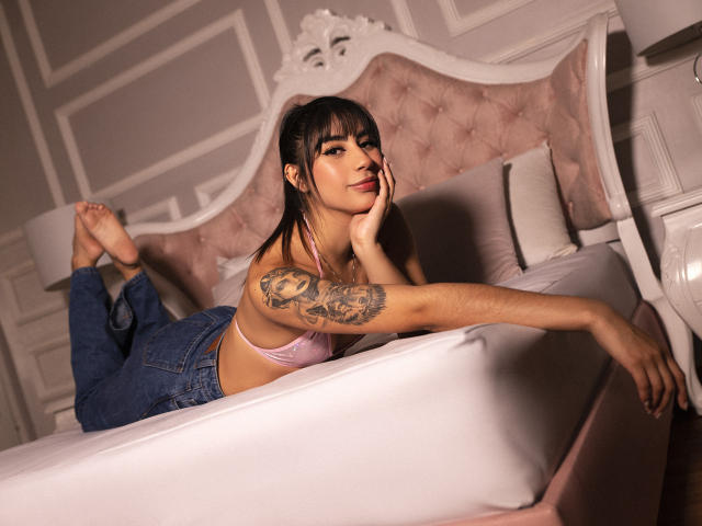 JessicaSoler, liggend op een bed, draagt een roze beha en blauwe jeans, heeft een tatoeage op haar rechterarm, haar benen zijn omhoog, haar handen liggen op het kussen, ze kijkt naar de camera, zachte, roze hoofdbord, casual, sexy..