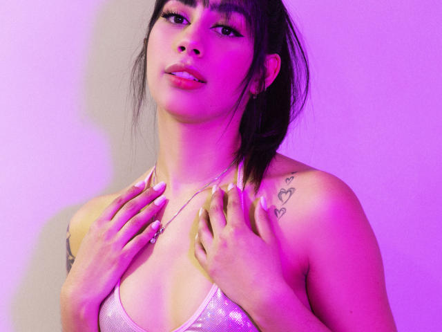 Jessica Soler, con iluminación rosa y púrpura, vistiendo un sujetador blanco brillante, con las manos en el pecho, tatuajes en el hombro, cabello negro, flequillo, mirando a la cámara, con los labios ligeramente entreabiertos..