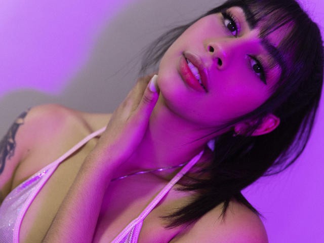 JessicaSoler has purple-tinted skin, dark bangs, and a hand on her neck, wearing a pink halter top, showing a tattoo on her left shoulder, with pink-lit face.