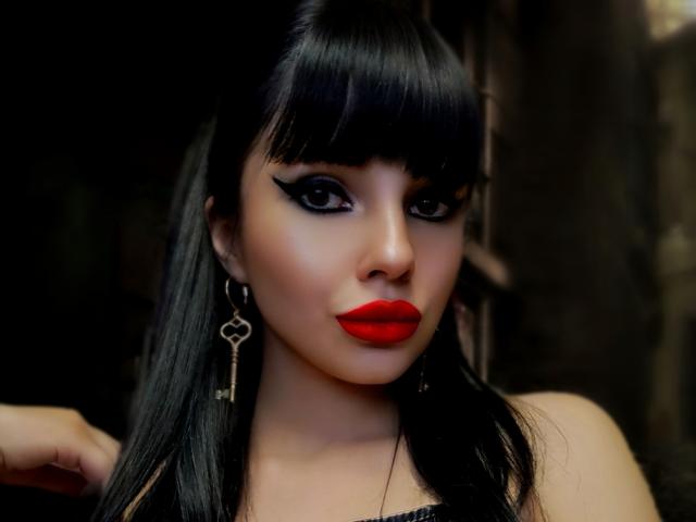 GoddessKaterina with black hair, red lipstick, thick eyeliner, and heart-shaped key earrings, looking seductive, her hand resting on her shoulder. Dark, moody background.