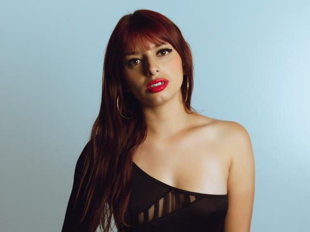 LeticiaFlorenz, met rood haar, oorringen en rode lippenstift, draagt een zwarte top met één schouder en uitsparingen, waardoor haar borsten zichtbaar zijn. Ze heeft een sexy, zelfverzekerde uitdrukking..