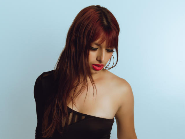 LeticiaFlorenz has red hair, looking down with pink lips, wearing a black, sheer, one-shoulder top, showing her bare, small boobs and a hint of her cleavage.