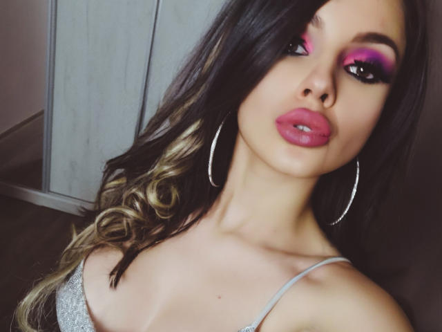 GoddessKaterina met roze oogmake-up, roze lippen en oorringen, draagt een zilveren top en laat haar borsten zien. Ze heeft lang, golvend bruin haar met blonde highlights..