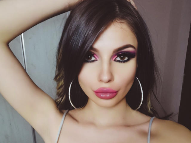 GoddessKaterina met donkerbruin haar, roze oogschaduw, groene ogen, roze lippenstift, oorringen en met haar armen omhoog, waarbij ze haar borsten en billen laat zien in een sexy, zelfverzekerde pose..