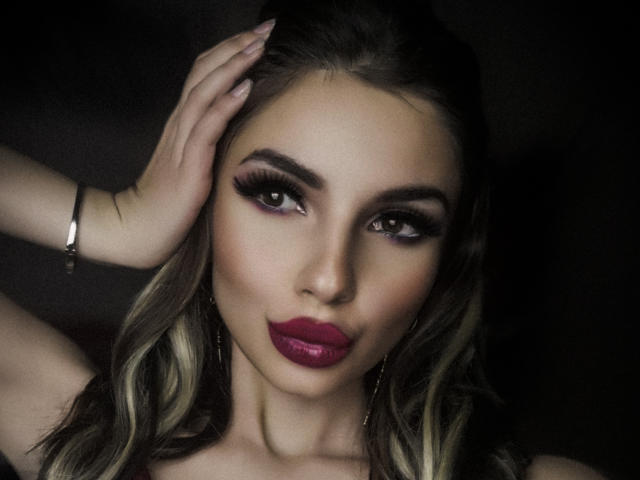 GoddessKaterina with pouty pink lips, smoky eye makeup, and wavy blonde hair, touching her head with a silver bracelet, looking seductively at the camera.