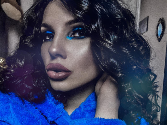 GoddessKaterina with dark, wavy hair, blue eye makeup, and glossy lips, wearing a fuzzy blue top, looking slightly to the side, with a hand near her head.