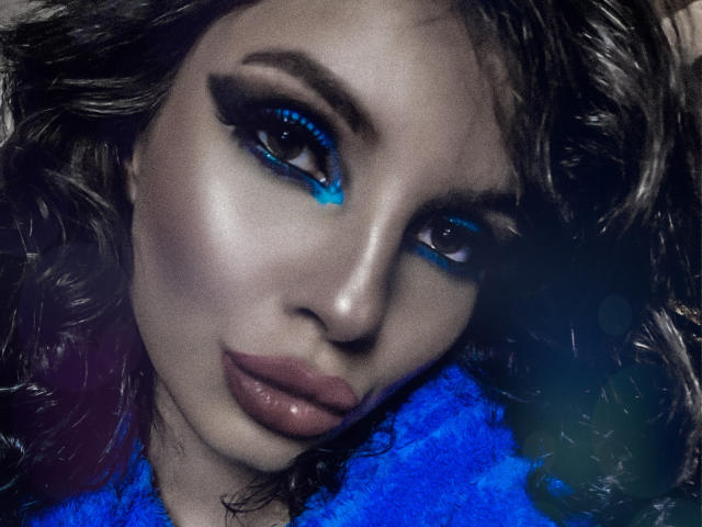 GoddessKaterina hat dunkles, welliges Haar, strahlenden blauen Lidschatten und volle, rosafarbene Lippen. Ihr attraktives Gesicht ist leicht geneigt, wodurch ihre glatte, ansprechende Haut zur Geltung kommt..