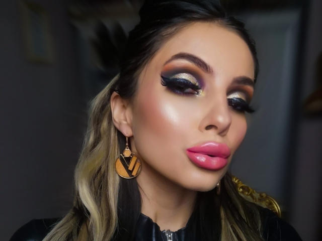 GoddessKaterina with shiny, purple, and gold eye makeup, pink lipstick, and black hair. She's wearing large, gold, V-shaped earrings, and has a sexy, makeup-sleeked face.