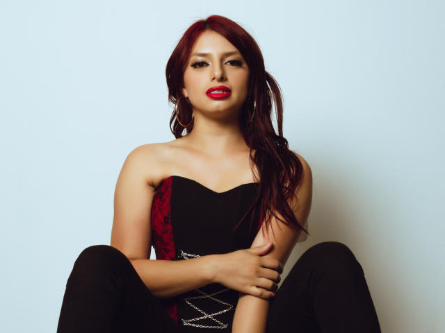 LeticiaFlorenz con i capelli rossi, vestito nero e rosso con lacci, seduta con le gambe divaricate, braccia incrociate sul petto, rossetto rosso, leggings neri, espressione sicura, sfondo bianco..