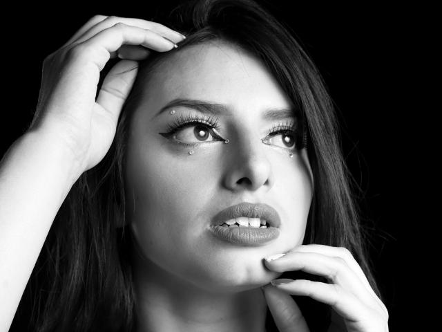 Il volto di LeticiaFlorenz in bianco e nero, mani sulla testa e sul mento, trucco occhi con piccoli strass, labbra leggermente aperte, sguardo rivolto verso l'alto, in monocromatico, espressione seria, il suo sedere non è visibile..