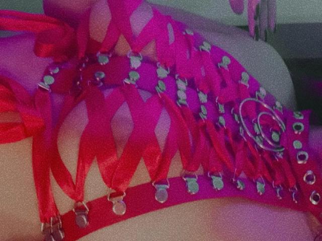 La lingerie rosa acceso di ShelbyShea, con spalline sottili, borchie e anelli, lascia intravedere i suoi seni attraverso motivi incrociati. La sua pelle chiara contrasta con il tessuto vivido e lucido..