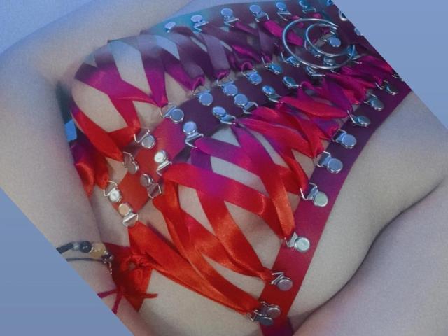ShelbyShea's red, strappy, cut-out top with metal rings and ribbons, showing her tits through the gaps, and a bracelet on her wrist, in a slightly angled shot.