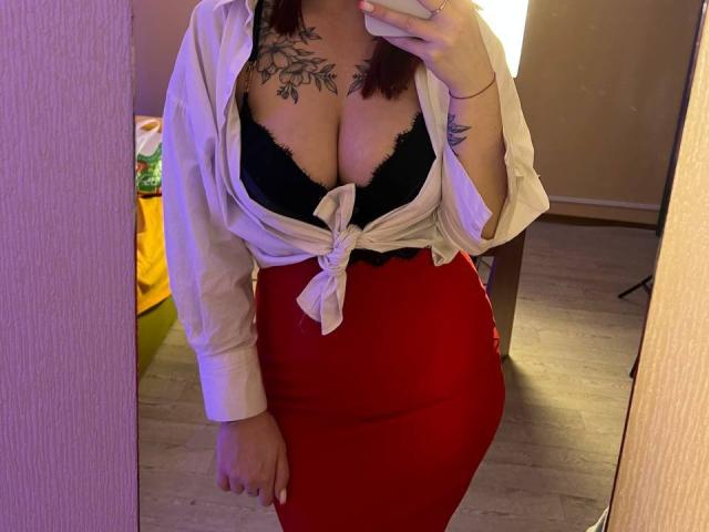 Innysik in a mirror selfie, wearing a tied white shirt revealing black lace boobs, red high-waist skirt, tattoo on chest, and wrist tattoo, holding a phone.