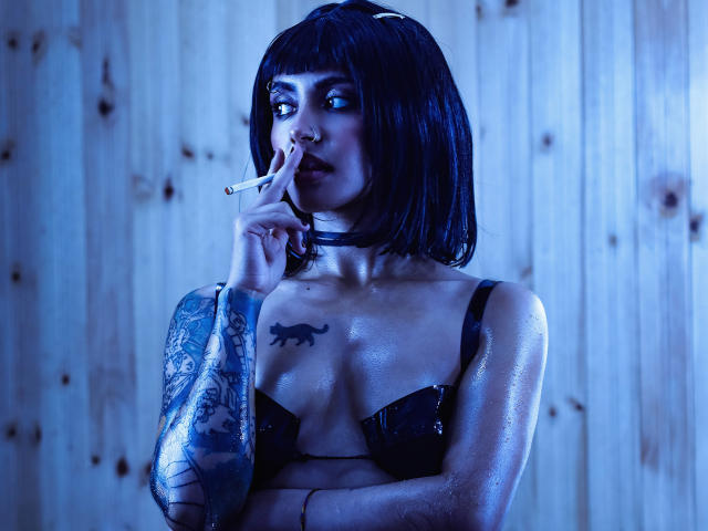 SerenaDeStark, avec sa coupe de cheveux courte noire, ses tatouages et sa peau humide, fume une cigarette, portant un soutien-gorge noir. Elle a un tatouage de panthère sur sa poitrine, devant un mur en bois éclairé en bleu..
