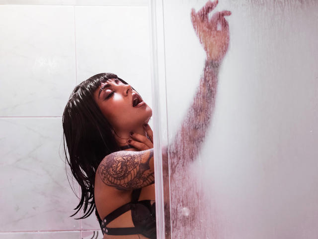 SerenaDeStark mit schwarzen Haaren, Tätowierungen am rechten Arm und dunklem Lippenstift, drückt ihre Hand und ihr Gesicht gegen eine beschlagene Glasduschtür und trägt schwarze Dessous..