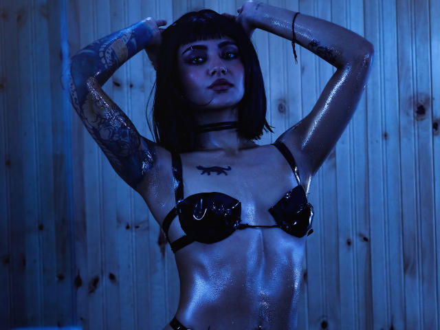 SerenaDeStark with wet skin, black glossy bra, arms raised, tattoos, choker, blue lighting, short black hair, serious expression, visible breast tattoos, wooden wall background.