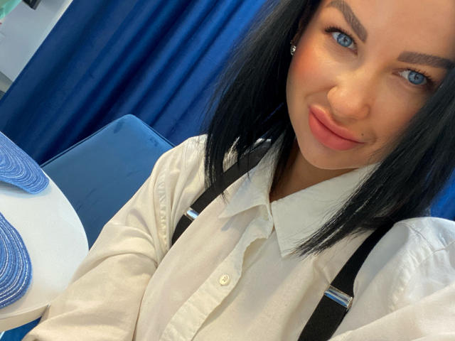 Kristen Stark mit blauen Augen, schwarzen Haaren und einem leichten Lächeln, trägt ein weißes Hemd mit schwarzen Hosenträgern und sitzt an einem blau bedeckten Tisch. Sie zeigt ein süßes Gesicht..