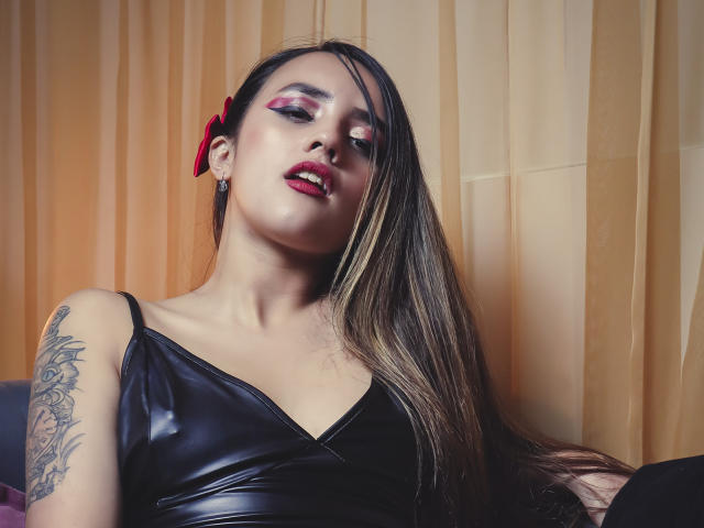 SabrinaCole met lang bruin haar, een rode strik, roze oogschaduw, een zwarte leren top, een tatoeage op de rechterarm, rode lippenstift, licht open lippen, naar beneden kijkend, gele gordijnen op de achtergrond..
