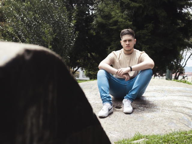 SebastiianMaxime è seduto a gambe chiuse su una roccia, indossa una maglietta beige, jeans blu e scarpe da ginnastica bianche, con le braccia appoggiate sulle ginocchia, in un parco pieno di erba..