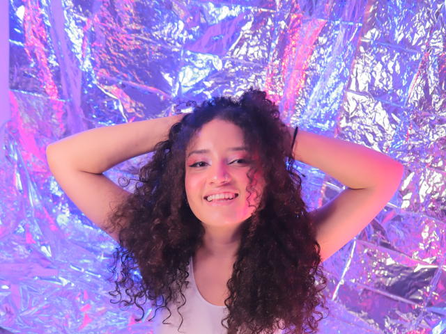 TracieLowe with curly black hair, smiling with hands behind her head, wearing a white tank top, purple and pink lighting on her face, and a shiny metallic background.